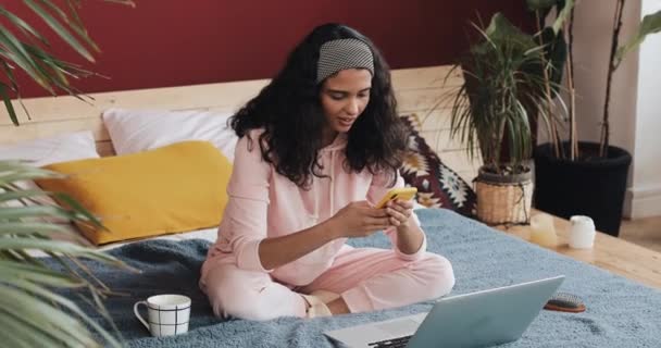 Chica afroamericana feliz charlando y mensajería de texto en el teléfono inteligente sentado en la cama en casa. Mujer joven recibe buenas noticias en el teléfono inteligente — Vídeo de stock