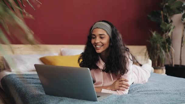 Happy african american girl lies on her belly on the bed by the window happily chatting with her friends on a video call on her laptop, slow motion — Stock Video