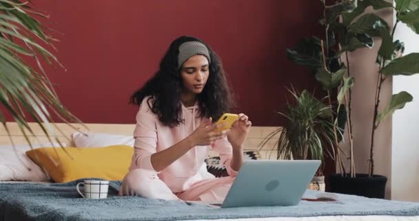 Ευτυχισμένος κορίτσι αφρικανική αμερικανική κουβεντιάζοντας και γραπτών μηνυμάτων στο smartphone που κάθεται στο κρεβάτι στο σπίτι. Νεαρή γυναίκα λαμβάνει καλές ειδήσεις για smartphone — Αρχείο Βίντεο