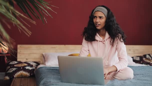 Hermosa chica vestida con pijama rosa usando computadora portátil sentada en la cama en casa. Mujer joven está sonriendo y charlando con amigos — Vídeo de stock