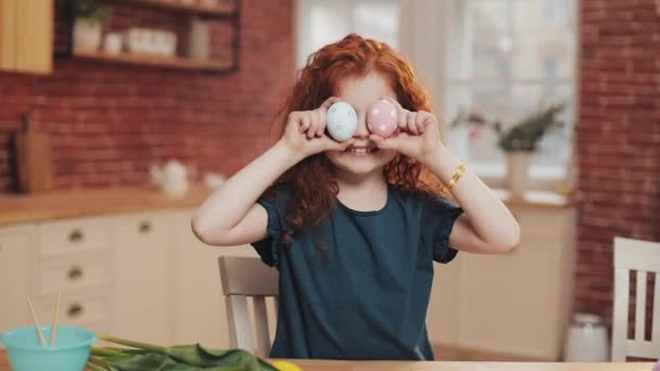 Portré vidám vörös hajú kislány játszik húsvéti tojás a konyhában háttérben. Éljenez és jól érzi magát a kamerában. Boldog Húsvétot!