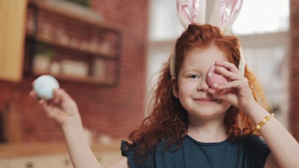 Portret van vrolijke roodharige kid beetje meisje met bunny oren met een easter egg op de achtergrond van de keuken. Ze is juichen en plezier op de camera. Vrolijk Pasen — Stockvideo