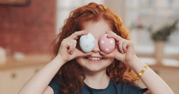 Mutfak arka planında paskalya yumurtasıyla oynayan neşeli kızıl saçlı küçük kız portresi. O, kameraya tezahürat yapıyor ve eğleniyor. Mutlu Paskalyalar