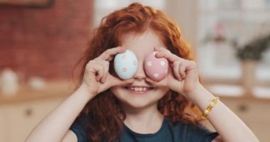 Mutfak arka planında paskalya yumurtasıyla oynayan neşeli kızıl saçlı küçük kız portresi. O, kameraya tezahürat yapıyor ve eğleniyor. Mutlu Paskalyalar