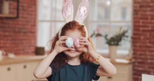 Retrato de niña pelirroja alegre con orejas de conejo con un huevo de Pascua en el fondo de la cocina. Ella está animando y divirtiéndose en la cámara. Feliz Pascua — Vídeos de Stock