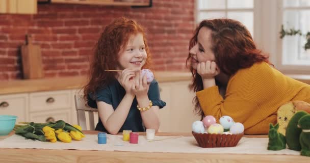 Η μαμά και η κόρη της ετοιμάζονται για το Πάσχα. Μικρό κορίτσι ζωγραφίζει πασχαλινά αυγά στη ζεστή κουζίνα. Χαμογελαστή μητέρα κοιτάζει την κόρη της. — Αρχείο Βίντεο
