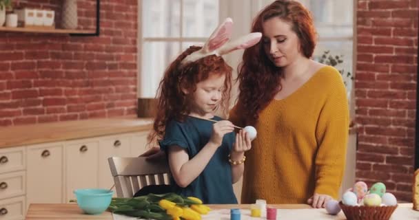 Счастливой Пасхи. Мама учит свою дочь рисовать яйца. Счастливая семья готовится к Пасхе. Маленькая девочка с кроличьими ушами — стоковое видео