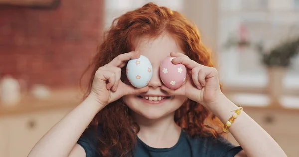 Mutfak arka planında paskalya yumurtasıyla oynayan neşeli kızıl saçlı küçük kız portresi. O, kameraya tezahürat yapıyor ve eğleniyor. Mutlu Paskalyalar