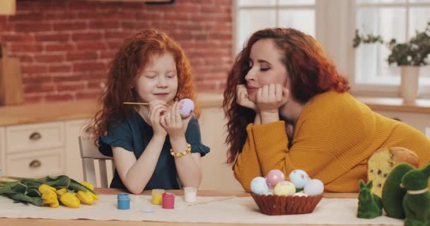Mamma e sua figlia si stanno preparando per Pasqua. Bambina che dipinge uova di Pasqua in cucina accogliente. Sorridente madre guarda sua figlia — Video Stock
