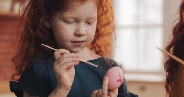 Alegre pelirroja niña con su madre pintando huevo de Pascua en el fondo de la cocina. Feliz Pascua. — Vídeo de stock