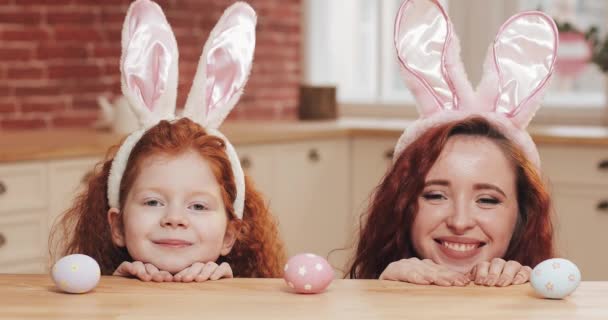 Портрет смішної мами та її дочки у вусі кролика з великодніми яйцями, які дивляться в камеру. щаслива сім'я розважається в затишній кухні. Щасливого Великодня — стокове відео