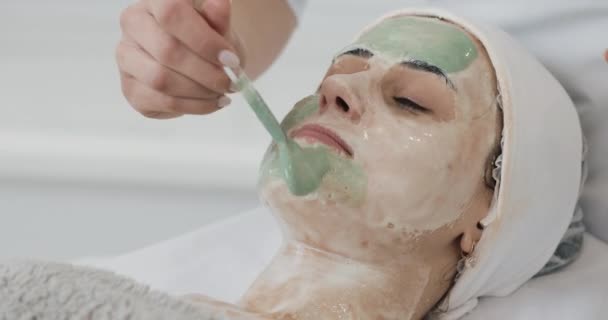 Piękna kobieta z twarzy maskę w salonie kosmetycznym. Kosmetolog stosowania zielonej twarzy maska do twarzy. Skóry procedur czyszczenia kosmetologii, zabiegi kosmetyczne spa — Wideo stockowe