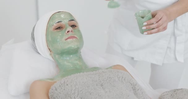 Belle jeune fille à l'esthéticienne fait les procédures de spa. Une main de cosmétologue applique un masque vert sur le visage des patients. Concept de soins corporels mode de vie sain — Video