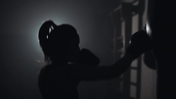Silhouet van jonge vrouw training aan een bokszak in een boxing club. Slow-motion. Achtergrond van de duisternis — Stockvideo