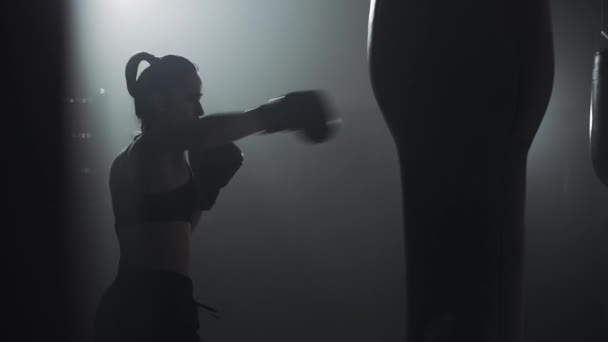 Silueta de mujer joven entrenando con un saco de boxeo en un club de boxeo. En cámara lenta. Fondo de oscuridad — Vídeos de Stock