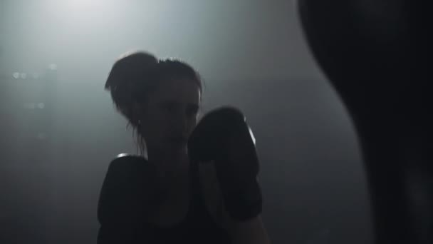 Силуэт молодой женщины, тренирующейся с боксерской грушей в боксерском клубе. Медленное движение. Тёмный фон — стоковое видео