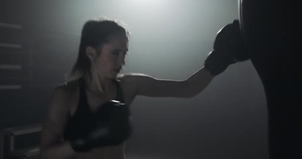 Bir boks kulübünde bir kum torbası ile eğitim genç kadın silüeti. Ağır çekim. Karanlık arka plan — Stok video