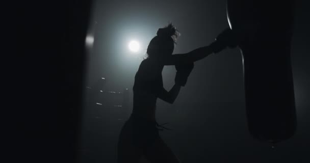 Silhueta de jovem treinando com um saco de perfuração em um clube de boxe. Movimento lento. Fundo de escuridão — Vídeo de Stock