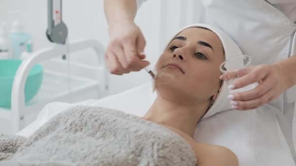 Mujer bonita joven acostada en el sofá en la clínica de cosmetología. Ella recibe máscara facial blanca en el salón de belleza del spa. Movimiento lento — Vídeo de stock