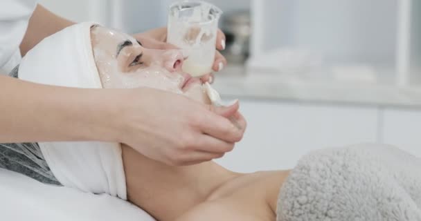 Jeune jolie femme allongée sur le canapé à la clinique de cosmétologie. Elle reçoit un masque facial blanc dans un salon de beauté spa — Video