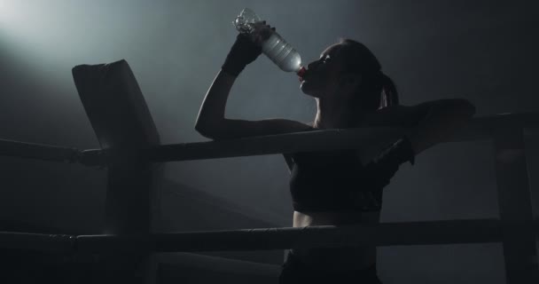 Mujer joven cansada bebiendo de la botella de agua después de entrenar en el anillo de boxeo oscuro con humo. Silueta. Concepto de boxeo — Vídeos de Stock