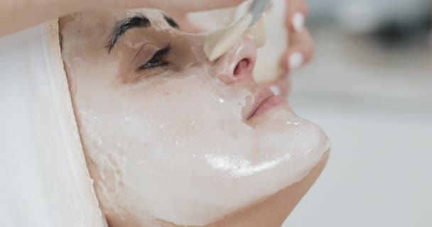 Mujer bonita joven acostada en el sofá en la clínica de cosmetología. Ella recibe máscara facial blanca en el salón de belleza del spa — Vídeo de stock