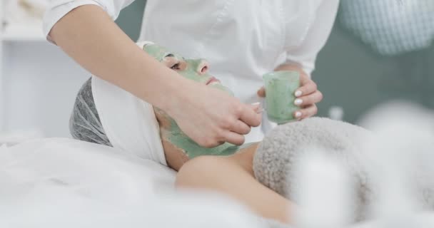 Piękna młoda dziewczyna w kosmetyczki robi zabiegi spa. Ręka kosmetologa stosuje zieloną maskę na twarzy pacjentów. Koncepcja pielęgnacji ciała zdrowy styl życia — Wideo stockowe