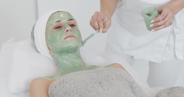 Hermosa joven en la esteticista hace los procedimientos de spa. Una mano del cosmetólogo aplica máscara verde en la cara de los pacientes. Concepto de cuidado corporal estilo de vida saludable — Vídeos de Stock