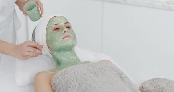 Vacker ung flicka på kosmetolog gör Spa förfaranden. En hand av kosmetolog applicerar grön mask på patientens ansikte. Begreppet kropps vård hälsosam livsstil — Stockvideo