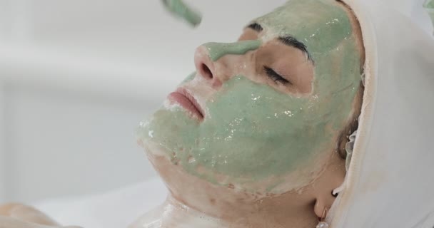 Piękna kobieta z twarzy maskę w salonie kosmetycznym. Kosmetolog stosowania zielonej twarzy maska do twarzy. Skóry procedur czyszczenia kosmetologii, zabiegi kosmetyczne spa — Wideo stockowe