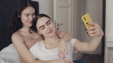 Mutlu lezbiyen çift yatağa sarılmak ve akıllı telefon ile birlikte selfie alarak kullanarak oturuyor. LGBT lezbiyen çift birlikte. Yavaş hareket.