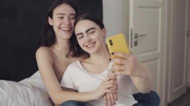 Mutlu lezbiyen çift yatağa sarılmak ve akıllı telefon ile birlikte selfie alarak kullanarak oturuyor. LGBT lezbiyen çift birlikte. Yavaş hareket.