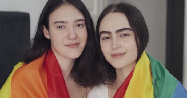 Portret van lesbische koppel met LGBT vlag op zoek naar de camera. Twee gelukkige vriendinnen poseren met Rainbow Flag at Home. — Stockvideo