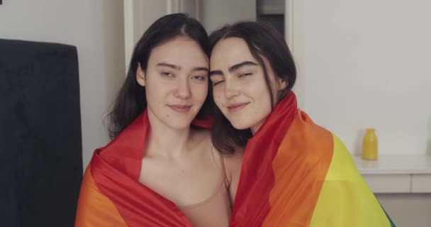 Retrato de pareja lesbiana con bandera LGBT mirando a la cámara. Dos amigas felices posan con la bandera del arco iris en casa . — Vídeo de stock