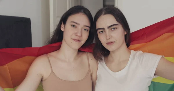 LGBT bayrağı kamera bakarak lezbiyen çift portre. İki mutlu kız evde gökkuşağı bayrağı ile poz.