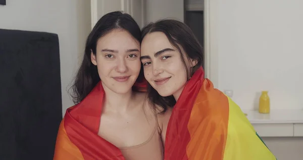 LGBT bayrağı kamera bakarak lezbiyen çift portre. İki mutlu kız evde gökkuşağı bayrağı ile poz.