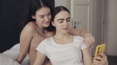 Mutlu lezbiyen çift yatağa sarılmak ve akıllı telefon ile birlikte selfie alarak kullanarak oturuyor. LGBT lezbiyen çift birlikte. Yavaş hareket.