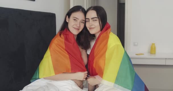 Портрет лесбийской пары с ЛГБТ-флагом, смотрящей в камеру. Две счастливые девушки позируют с радужным флагом дома . — стоковое видео