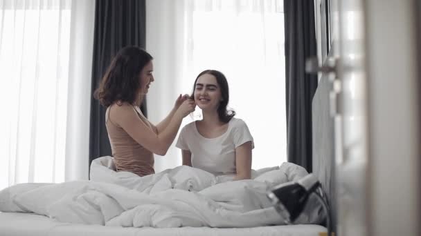 Lesbisk kvinna kamning hår hennes flickvän. Lycklig lesbisk par sitter i sängen hemma. Romantiskt Relations koncept. Slow motion. — Stockvideo