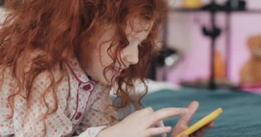 Sarı akıllı telefon ile Komik kızıl saçlı küçük kız yatakta yatıyor ve akıllı telefon kullanarak. İletişim, oyun oynama, uygulama.