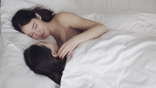 Pareja de lesbianas abrazándose y sonriendo mientras yacen juntas en la cama en casa. Jóvenes lesbianas besos y abrazos después de despertar — Vídeo de stock