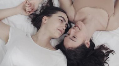 Mutlu lezbiyen kızlar yatakta yatıyorlar, gülümsüyorlar ve birbirlerinde eğleniyorlar. LGBT aile kavramı, yavaş çekim. Üstte görüntü.