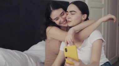 Mutlu lezbiyen çift yatağa sarılmak ve akıllı telefon ile birlikte selfie alarak kullanarak oturuyor. LGBT lezbiyen çift birlikte. Yavaş hareket.