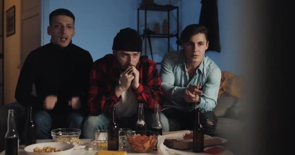 Tres amigos viendo el deporte en la televisión, preocupándose por la pérdida del equipo. Chicos sentados en el sofá en casa con cerveza y aperitivos. Emoción, verdaderos fans — Vídeos de Stock