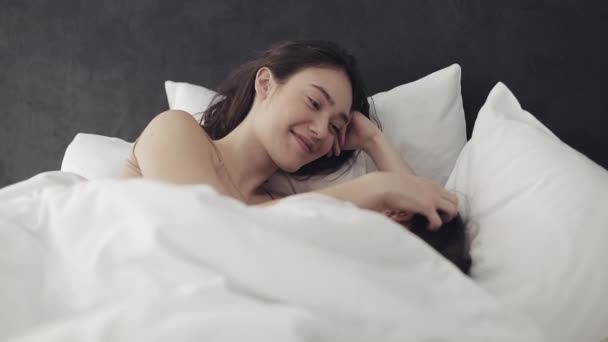 Лесбиянки обнимаются и улыбаются, лежа вместе в постели дома. Молодые лесбиянки целуются и обнимаются после пробуждения — стоковое видео