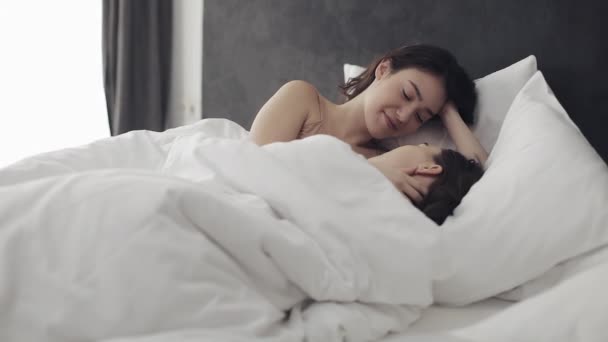 Pareja de lesbianas abrazándose y sonriendo mientras yacen juntas en la cama en casa. Jóvenes lesbianas besos y abrazos después de despertar — Vídeos de Stock