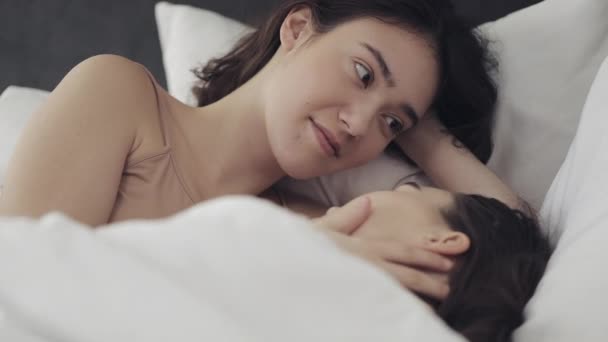 レズビアンのカップルは、自宅でベッドに一緒に横たわっている間、抱擁し、笑顔。若いレズビアンは目を覚ます後にキスと抱擁 — ストック動画