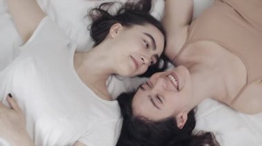 Mutlu lezbiyen kızlar yatakta yatıyorlar, gülümsüyorlar ve birbirlerinde eğleniyorlar. LGBT aile kavramı, yavaş çekim. Üstte görüntü.