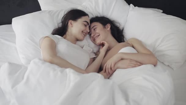 Лесбиянки обнимаются и улыбаются, лежа вместе в постели дома. Молодые лесбиянки целуются и обнимаются после пробуждения. Вид сверху . — стоковое видео