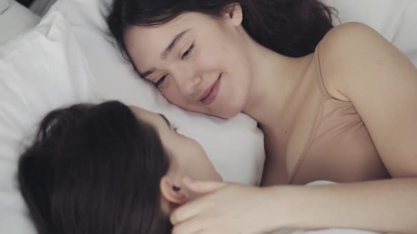 Pareja de lesbianas abrazándose y sonriendo mientras yacen juntas en la cama en casa. Jóvenes lesbianas besos y abrazos después de despertar — Vídeo de stock